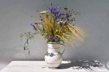A bouquet of meadow wild flowers in a white jug on a wooden white table, in the background a white wall with a shadow
