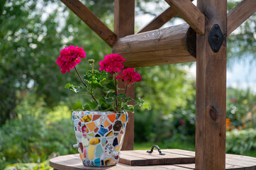 Homemade mosaic flower pot with porcelain pieces. Recycling, broken tableware, ceramics, crafts, mosaics. The vase stands on top of the well. Red geranium blooms.