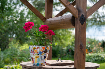 Homemade mosaic flower pot with porcelain pieces. Recycling, broken tableware, ceramics, crafts, mosaics. The vase stands on top of the well. Red geranium blooms.