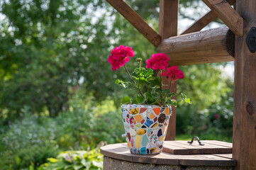 Homemade mosaic flower pot with porcelain pieces. Recycling, broken tableware, ceramics, crafts, mosaics. The vase stands on top of the well. Red geranium blooms.
