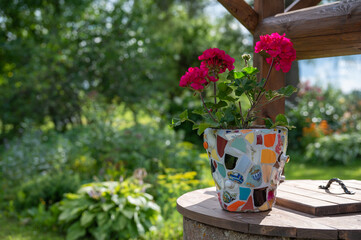 Homemade mosaic flower pot with porcelain pieces. Recycling, broken tableware, ceramics, crafts, mosaics. The vase stands on top of the well. Red geranium blooms.