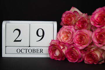 October 29 wooden calendar, white on a black background, pink roses lie nearby. Postcard with copy space. The concept of a holiday, congratulation, invitation, party, announcement, vacation, promotion