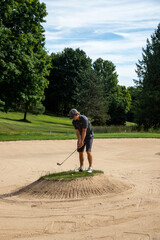 Male golfer in gray with ball in sand trap, bunker shot, end in sight, overcoming obstacles, challenging shot, target in sight, island in bunker