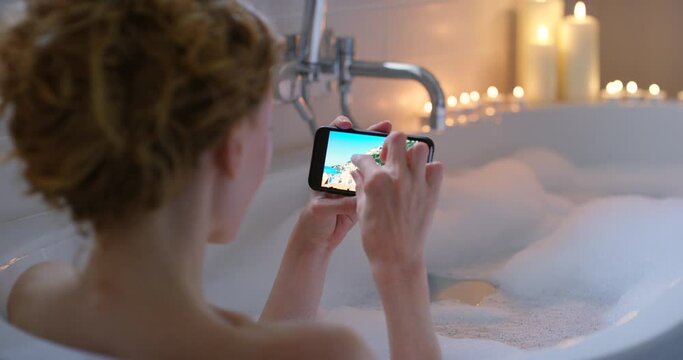 A woman relaxing in her bubble bathtub and looking at holiday vacation getaway photos on social media. Modern carefree female enjoying a cozy ambience filled night in her bathroom at home alone
