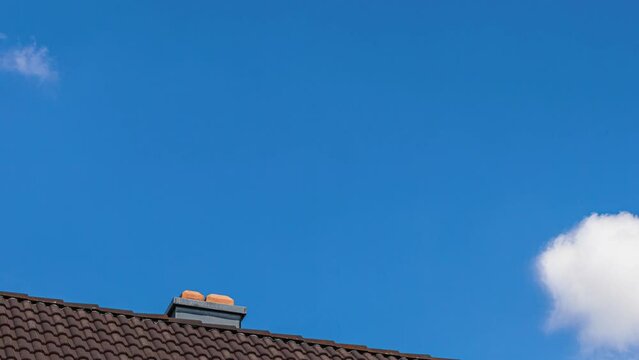 Daytime time lapse of clouds floating across the blue sky over the roof of a house with a chimney
