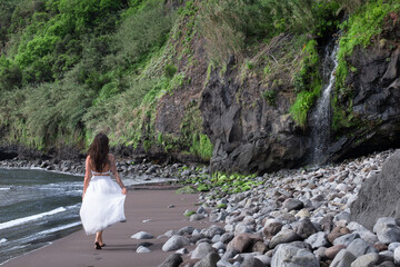 Frau mit weißen Kleid läuft am atemberaubenden schwarzen Sandstrand auf Madeira entlang, mit...