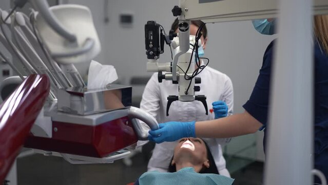 Dental microscope with professional doctor examining patient and assistant moving tools closer in slow motion. High-quality equipment and expert dentist treating Caucasian woman in hospital