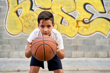 A cute young boy plays basketball on the street playground in summer. Teenager in a white t-shirt with orange basketball ball outside. Hobby, active lifestyle, sports activity for kids