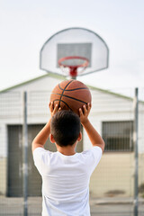A cute young boy plays basketball on the street playground in summer. Teenager in a white t-shirt...