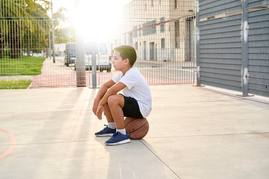 A Cute Young Boy Plays Basketball On The Street Playground In Summer. Teenager In A White T-shirt With Orange Basketball Ball Outside. Hobby, Active Lifestyle, Sports Activity For Kids