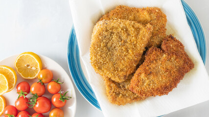 Crispy pan fried breaded pork chops served with lemon and fresh tomatoes close up on a plate on light grey background