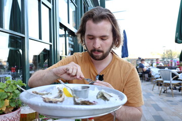 young white male with long hair and beard, wearing mustard t-short, sitting outside in the restaurant eating fresh oysters with his hands