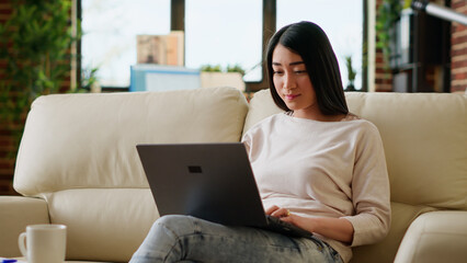 Smiling office worker at home working remotely on laptop while sitting on sofa in living room. Young adult person doing remote work on modern portable computer while at home.