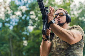 Caucasian bearded man in safety goggles and headphones wearing camo t-shirt reloading submachine gun. Outdoor horizontal shot. High quality photo