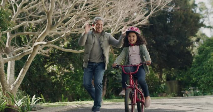 Grandfather teaching child to cycle on her first bike ride, happy child outdoors learning on a bicycle in a street. Smiling, clapping and proud grandpa having a bonding moment with his grandchild.