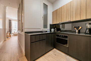 Open kitchen with low black and high wooden cabinets, integrated appliances and a corridor leading to a living room with a balcony