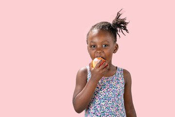 ritratto in studio isolato con sfondo rosa di bambina nera che mangia una mela  - obrazy, fototapety, plakaty