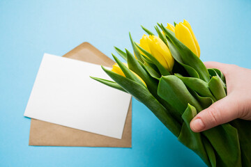 Hand holds a bouquet of yellow tulips. Empty letter for text on blue background. Gift and Greetings