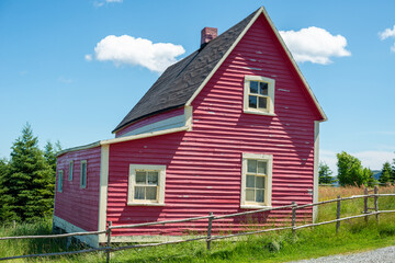 Obraz premium A view of a vintage cottage under blue sky and clouds. The small red wooden building has multiple double hung windows with a black roof. There's an old log fence, green grass, and a gravel path. 