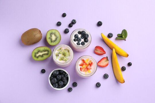 Jars of fresh yogurt and different fruits on lilac background, flat lay