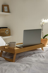 Wooden tray with modern laptop and cup of aromatic drink on bed indoors