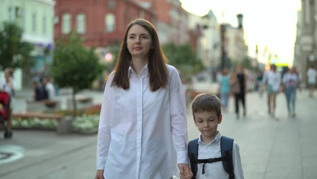 Happy Mom And Son Holding Hands After The Lessons In School. Mother Picks Up A Schoolboy From School And Walks Home Down The Street Against The Background Of Defocused People