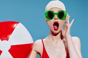 A surprised shocked lady in glasses a swimming cap a red swimsuit with her mouth open holds a swimming ball