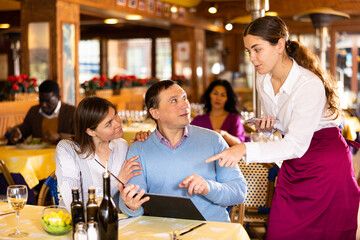 Polite young waitress using smartphone to take order in restaurant, helping couple choose dishes from menu..