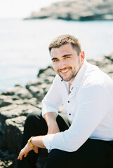 Fototapeta na wymiar Smiling man in a white shirt sits on a stone by the sea