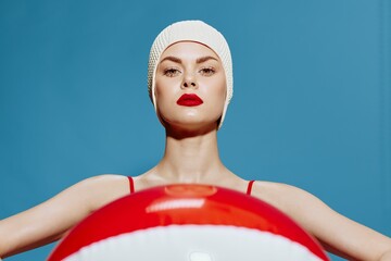 Woman in a stylish swimsuit swim cap stretches a striped ball forward while looking at the camera...