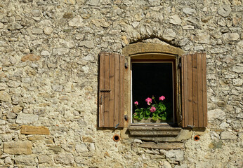 Window with pink geranium flowers and open wooden shutters in a stone wall. From the hamlet of La Chaudiere, in the Rhone-Alpes region of France.