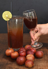 Hand clicking glass with plum wine cocktail on wooden table with plums and black background
