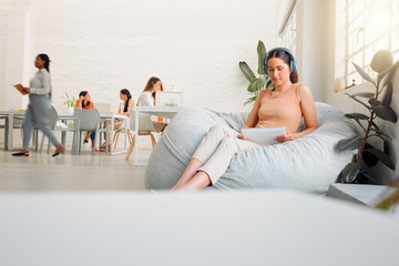 Creative intern browsing online with a tablet, listening to music on wireless headphones. Female on a break sitting on bean bags in a casual office space to improve creativity, work and mindfulness