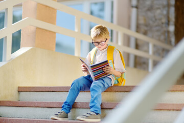 Smart little child sitting and reading on the stairs of school building. Quality education for children. Portrait of funny nerd schoolboy with big glasses. Vision problems. Back to school concept. - obrazy, fototapety, plakaty