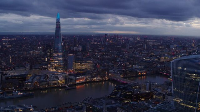 Panoramic Drone establishing shot of illuminated Central London at dusk. Iconic landmarks like the London Bridge, The Shard and the River Thames standing out from the rest of the city.