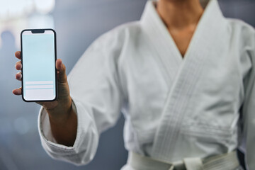 . Blank phone screen and copyspace with a karate student holding the mobile display indoors. Sport person showing a copy space announcing a sale, marketing or social media advertising content. © Delcio F/peopleimages.com