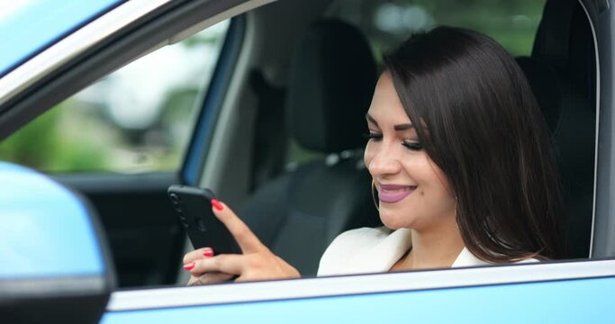 Young woman driver types messages to friend on black smartphone smiling. Stylish businesswoman with perfect makeup sits in car texting friend closeup