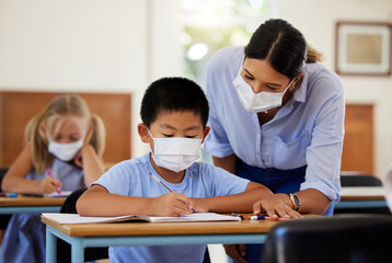 Covid, education and learning with a teacher wearing a mask and helping a male student in class during school. Young boy studying in a classroom with help from an educator while sitting at his desk.