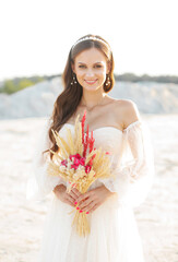 Fototapeta na wymiar A pretty brunette girl in a white dress stands casually with a bouquet of dry flowers on the background of a white sand quarry, wedding concept