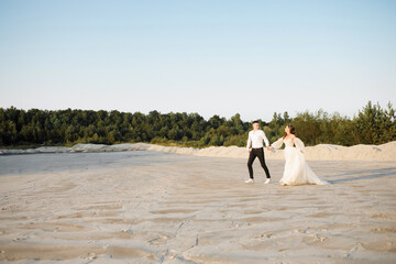 Fototapeta na wymiar Cute newlyweds are running on the sand against the background of the forest, holding hands and looking at the sun