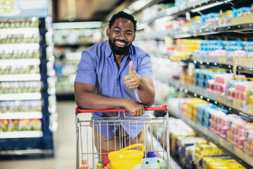 Black Male Buyer Shopping Groceries In Supermarket Standing With Shop Cart Indoors Showing Thumb Up.