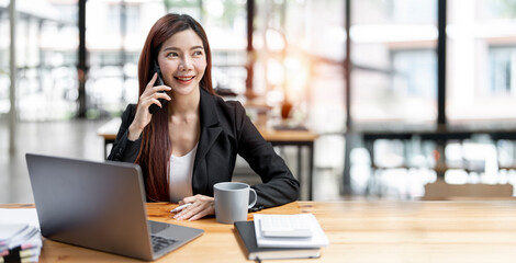 Fototapeta premium Happy businesswoman sitting at desk behind her laptop and talking with somebody on her mobile phone while working at office.