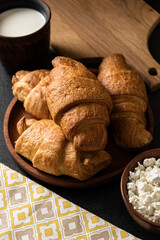 Cozy breakfast with croissants, cottage cheese and milk on a dark background with textiles, wooden and clay dishes. French pastries. Rustic breakfast.