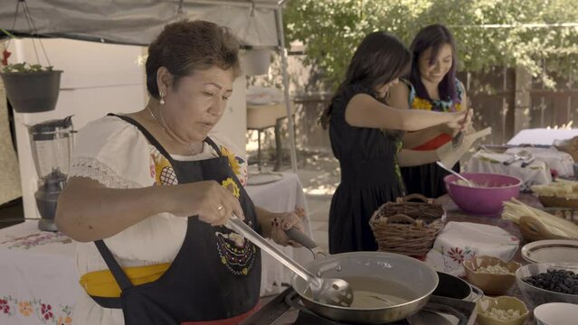 Indigenous woman takes plums out of a pan with oil as her twin daughters make tamales beside her. 