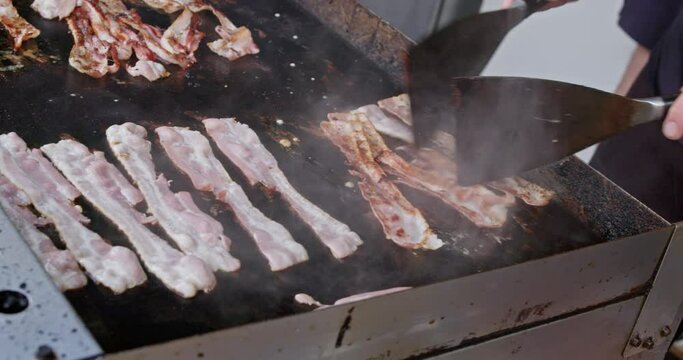A street food vendor cooks and flips bacon rashers on a gas griddle plate at a street food market.
