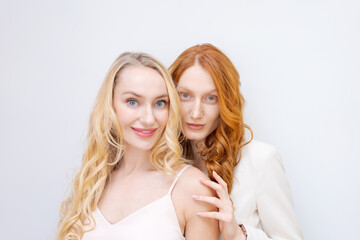 Close-up fashion portrait couple of two pretty women, best friends smiling on light background. Variable wavy hairstyle. Blonde and redhead posing
