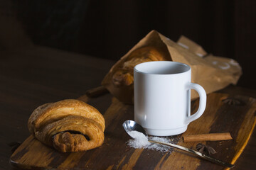 Baked fresh croissants with chocolate in the kitchen interior on a wooden dividing board on a dark background. Nearby is a white cup of coffee with sugar and cinnamon. Photos of food for cafes.