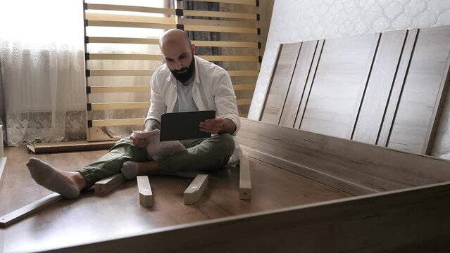 Bearded man with tattoo on the hand with instruction in the tablet for assembling furniture is going to assemble a wooden bed in a bedroom. Heavy assembly of home furniture by yourself.
