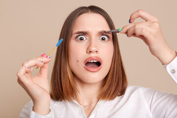 Portrait of shocked scared female in beauty salon holding eyebrow brush and tweezers, looking at camera with big eyes, being shocked after plucking eyebrow procedure.