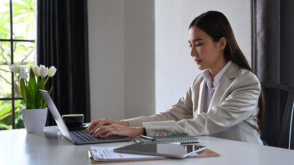 Concentrated asian female accountant working and checking online information on laptop at her workplace